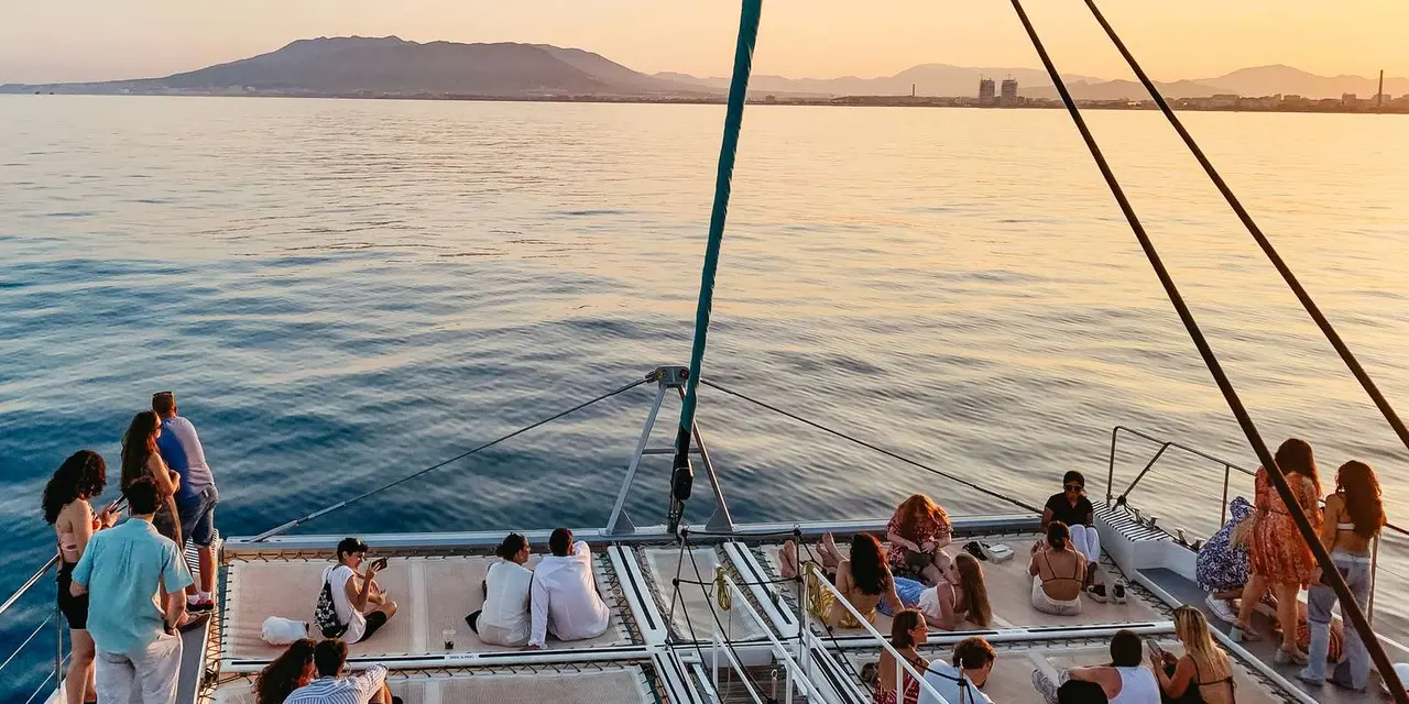 A catamaran's rigging and sails silhouetted against a calm sea at sunset, with mountains on the horizon
