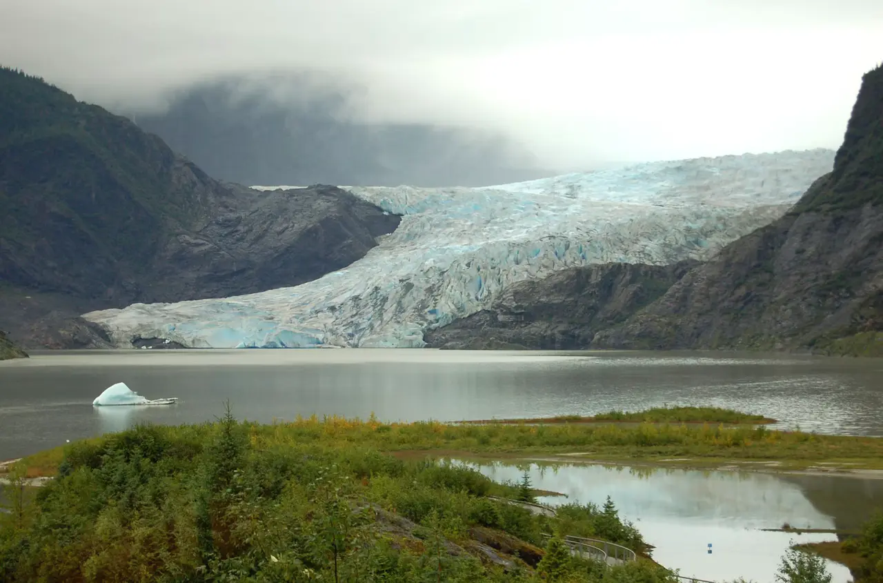 Mendenhall Glacier's blue-white ice flowing between dark mountain walls into a grey lake, with a small iceberg floating near the shore and low cloud above