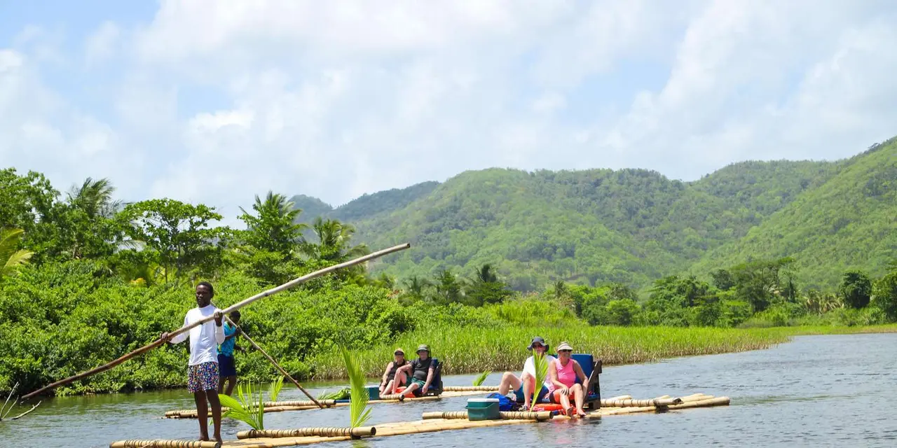 A guide poling a bamboo raft with passengers along a river, with green hills behind