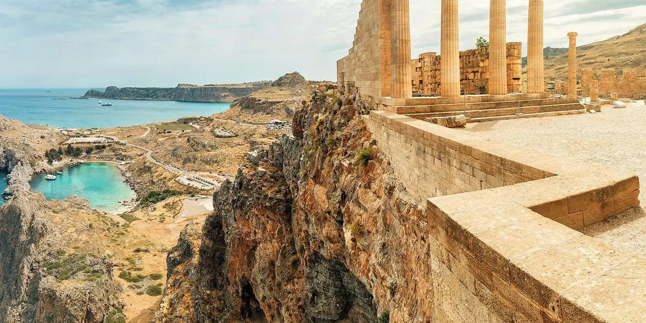 The columns and walls of the Lindos Acropolis on a rocky cliff above a bay with a small beach
