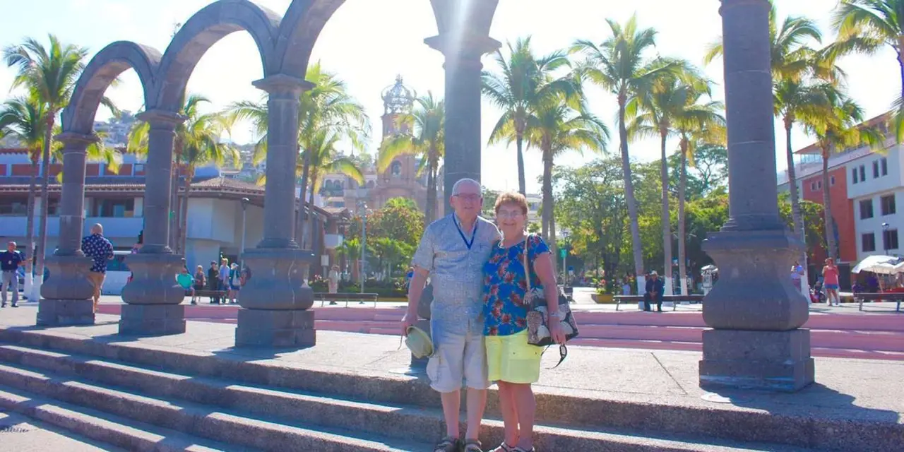 A couple standing at the foot of stone arches with palm trees and a church tower behind them on a sunny plaza