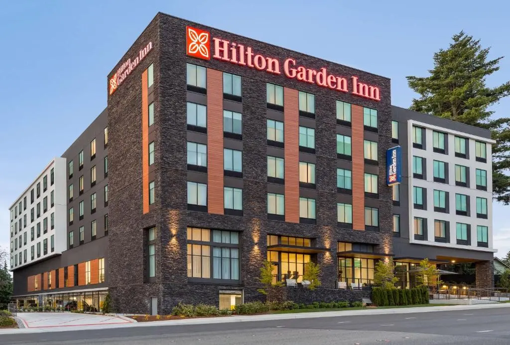 The dark-brick Hilton Garden Inn Seattle Airport with its red sign on the corner at dusk