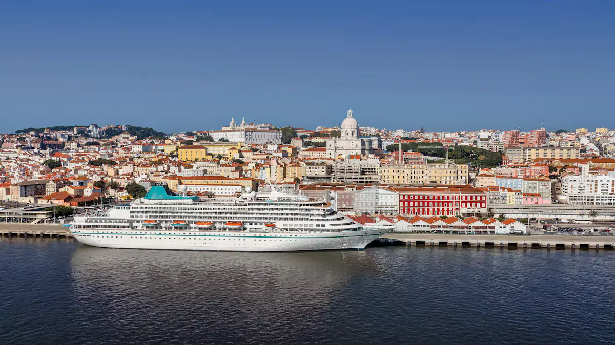 A white cruise ship moored on the Tagus at Lisbon's eastern waterfront, with the red Santa Apolónia station front, Alfama's pastel houses and the National Pantheon dome behind under a clear blue sky