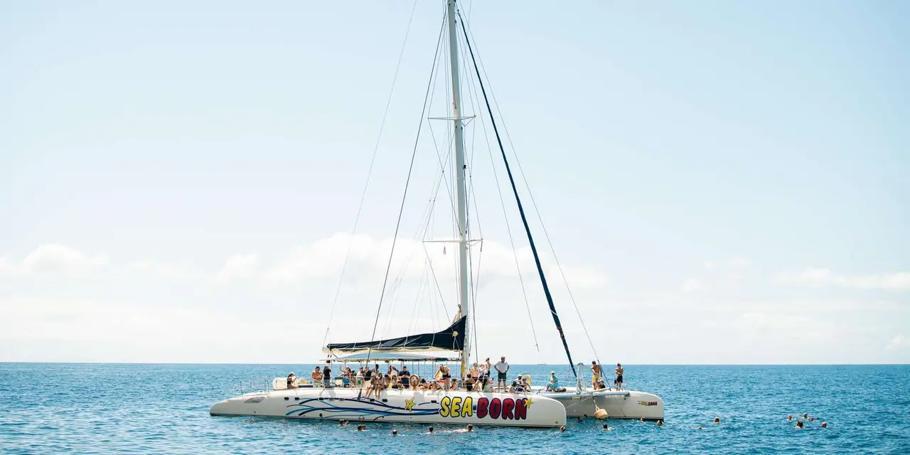 A white sailing catamaran with a tall mast and passengers on deck on calm, pale blue water