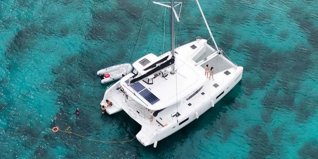 A white catamaran with a tall mast seen from above on clear turquoise water, a swimmer beside it