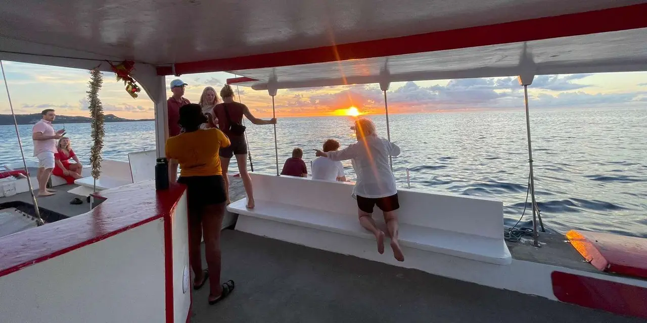 Guests on the shaded deck of a sailing boat watching the sun set over the sea