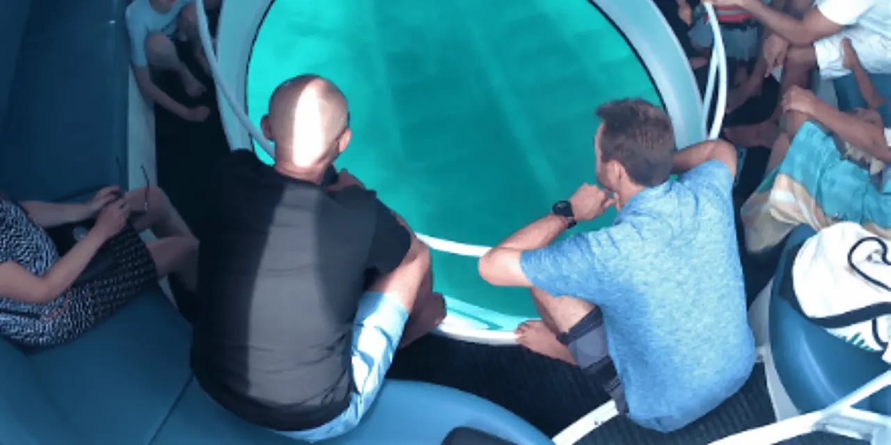 Passengers seated around a large round glass window in the floor of a boat, looking down into blue water