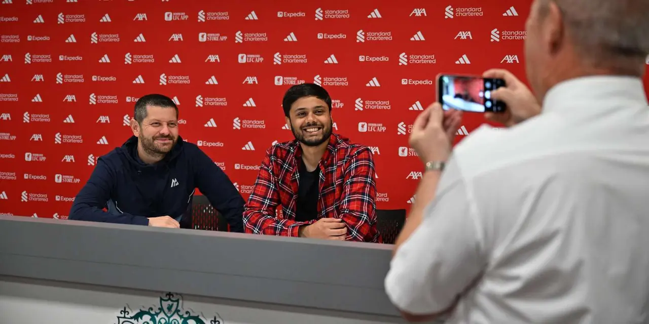 Two visitors smiling for a photo in front of the red sponsor backdrop in Liverpool FC's press-conference room while a third person takes their picture with a phone