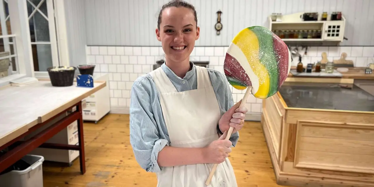 A smiling cook in an apron holding a large striped lollipop in a white-tiled sweet shop