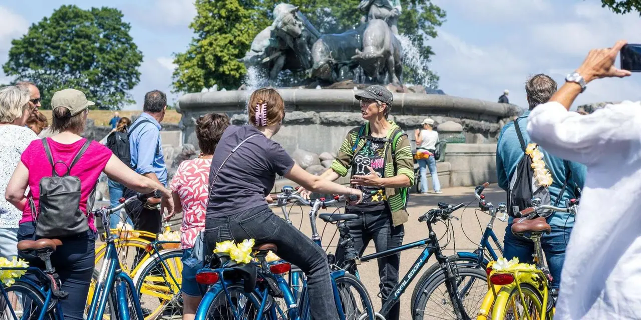 A guide talking to a group on bikes in front of the bronze Gefion Fountain, with the oxen and goddess figures behind them under a blue sky