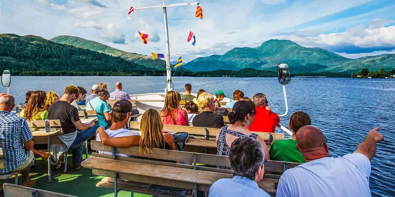 Passengers at picnic tables on the open deck of a boat strung with signal flags, green hills along the loch behind