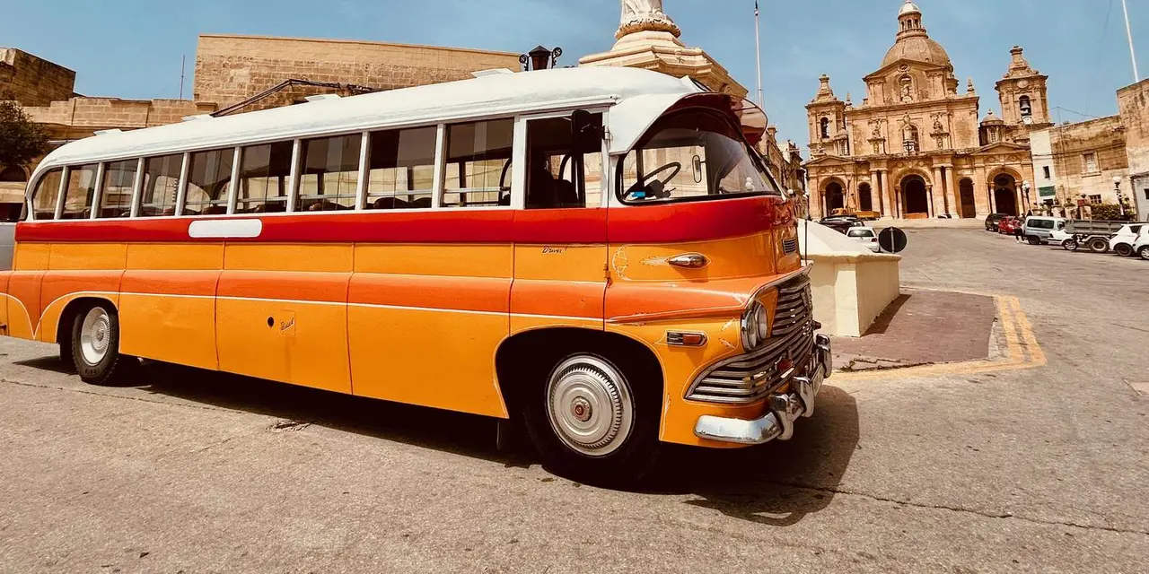 An orange-and-cream vintage Maltese bus parked beside a church statue