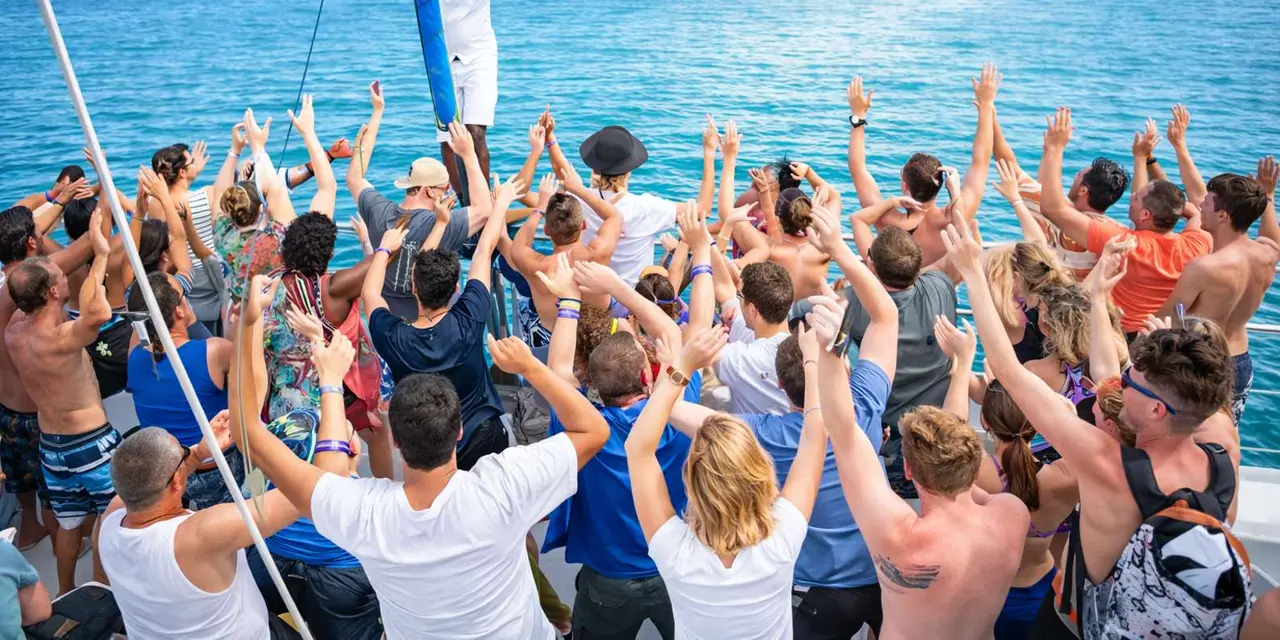 A crowd of passengers with arms raised on a boat deck in front of blue water