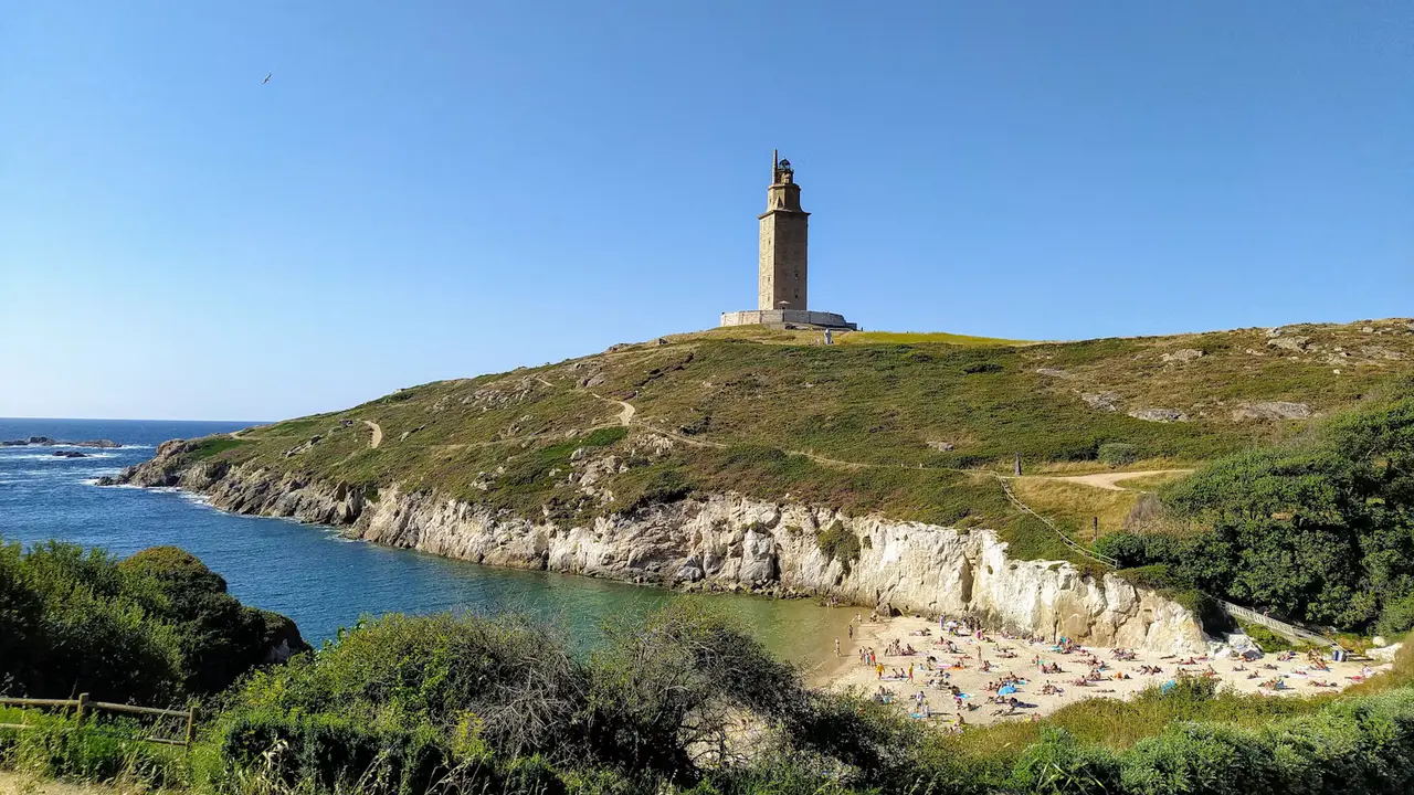 The Tower of Hercules standing on a green headland above a small sandy cove and blue water in A Coruña, under a clear blue sky