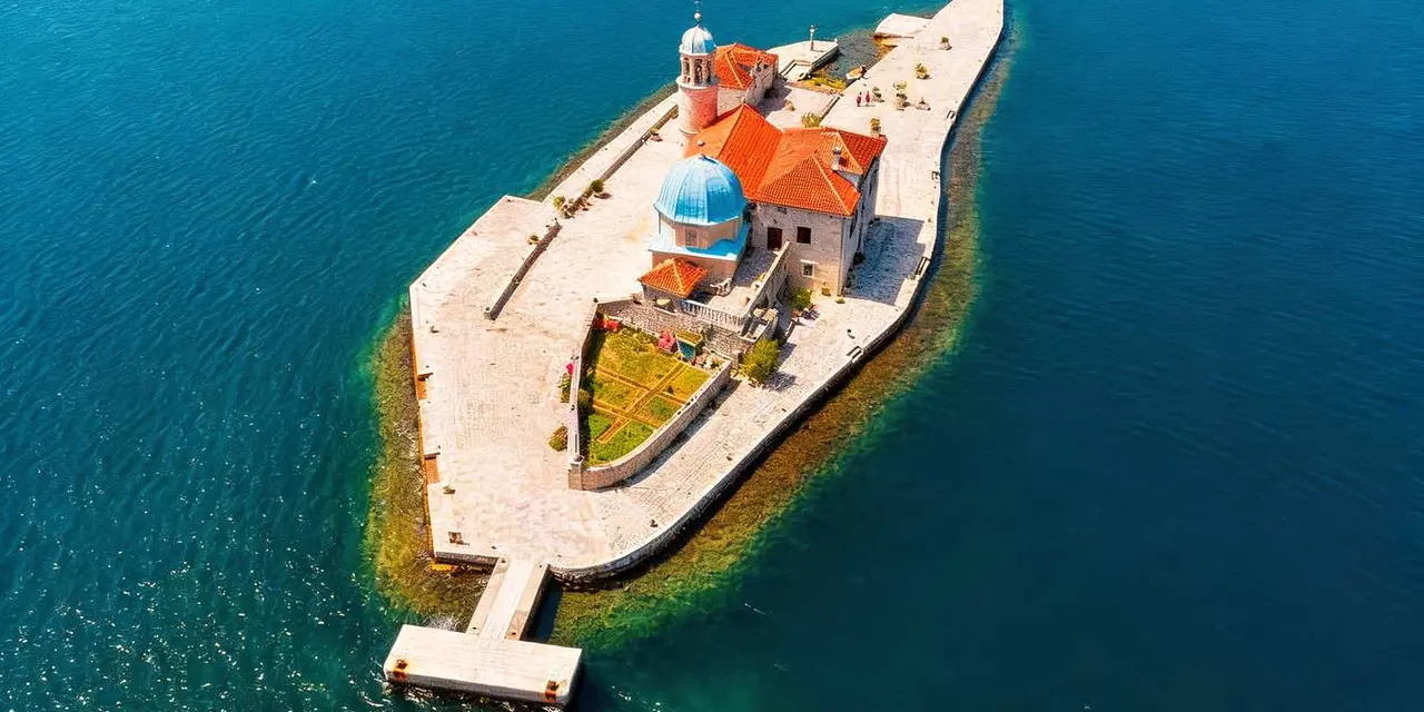 An aerial view of Our Lady of the Rocks, a small island with a blue-domed church and red roofs in deep blue water