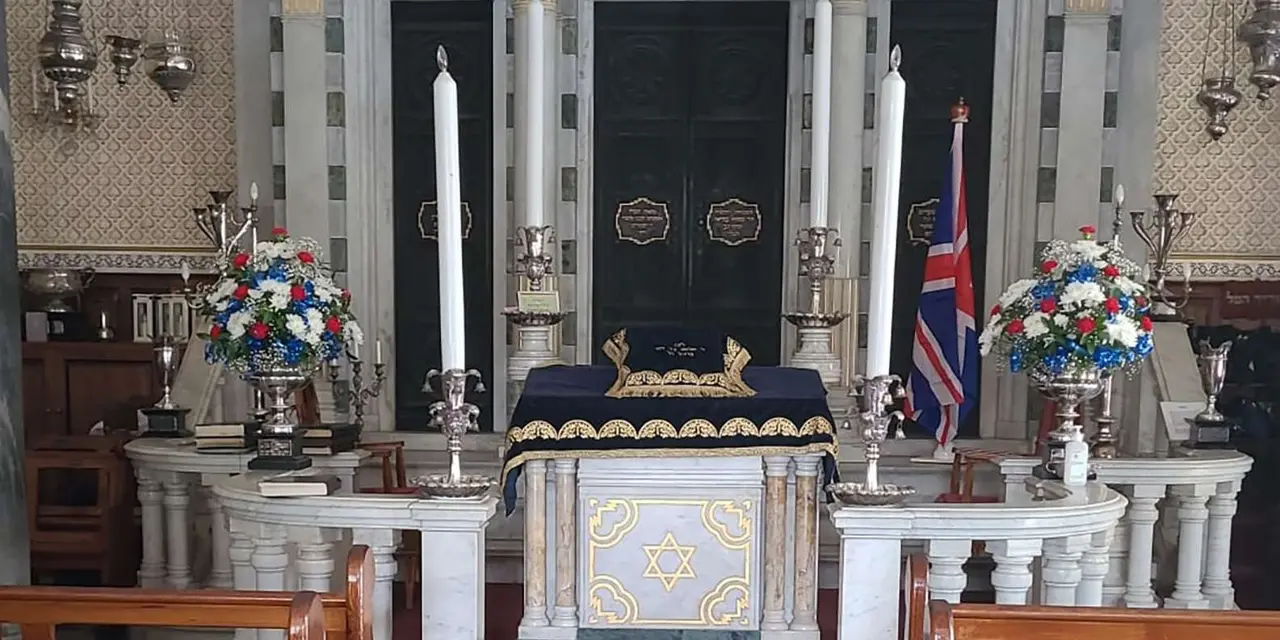 The interior of a Gibraltar synagogue with tall white candles, flowers and a Union flag beside the ark