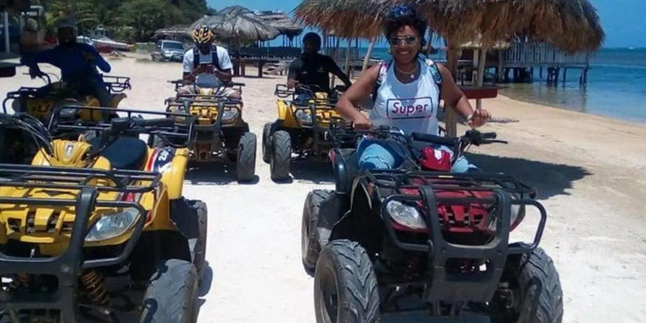 Visitors in helmets sit on yellow and red ATVs parked on a sandy track beside thatched beach huts