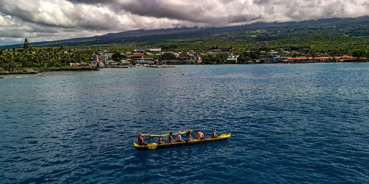 A crew paddling a long outrigger canoe across calm blue water in front of Kailua-Kona's shoreline