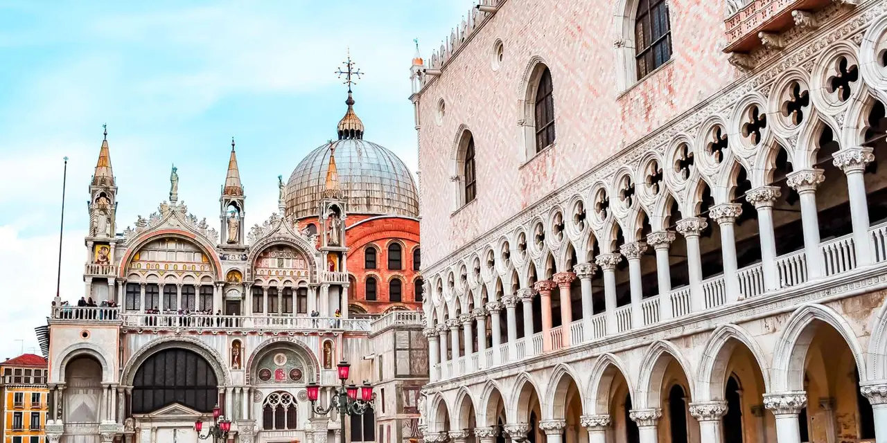 The pink-and-white Gothic arcades of the Doge's Palace with the domes of St. Mark's Basilica behind them under a blue sky