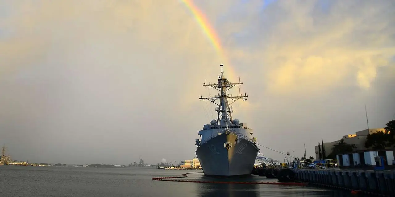 A grey Navy warship moored at a pier beneath a rainbow and a cloudy evening sky