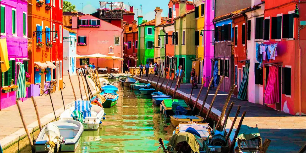 A canal on Burano lined with houses painted pink, orange and green, with small boats moored along the bank