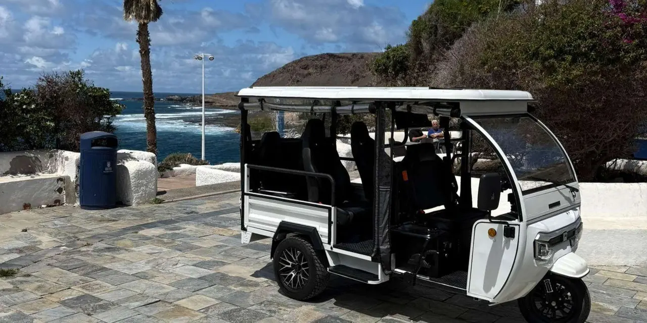 A white electric tuk-tuk parked on a paved seafront with palms and the sea behind