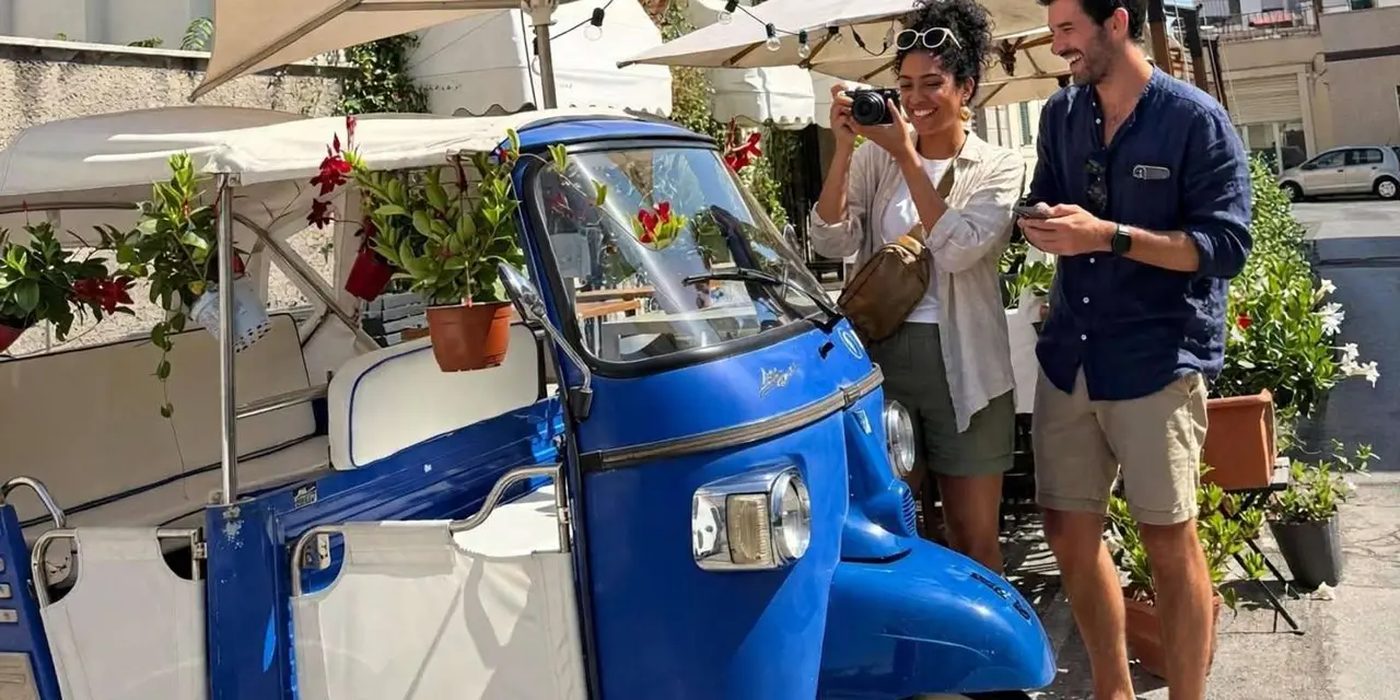 A blue three-wheeled van decorated with potted plants parked in a market street, with a guide and a guest taking photos beside it