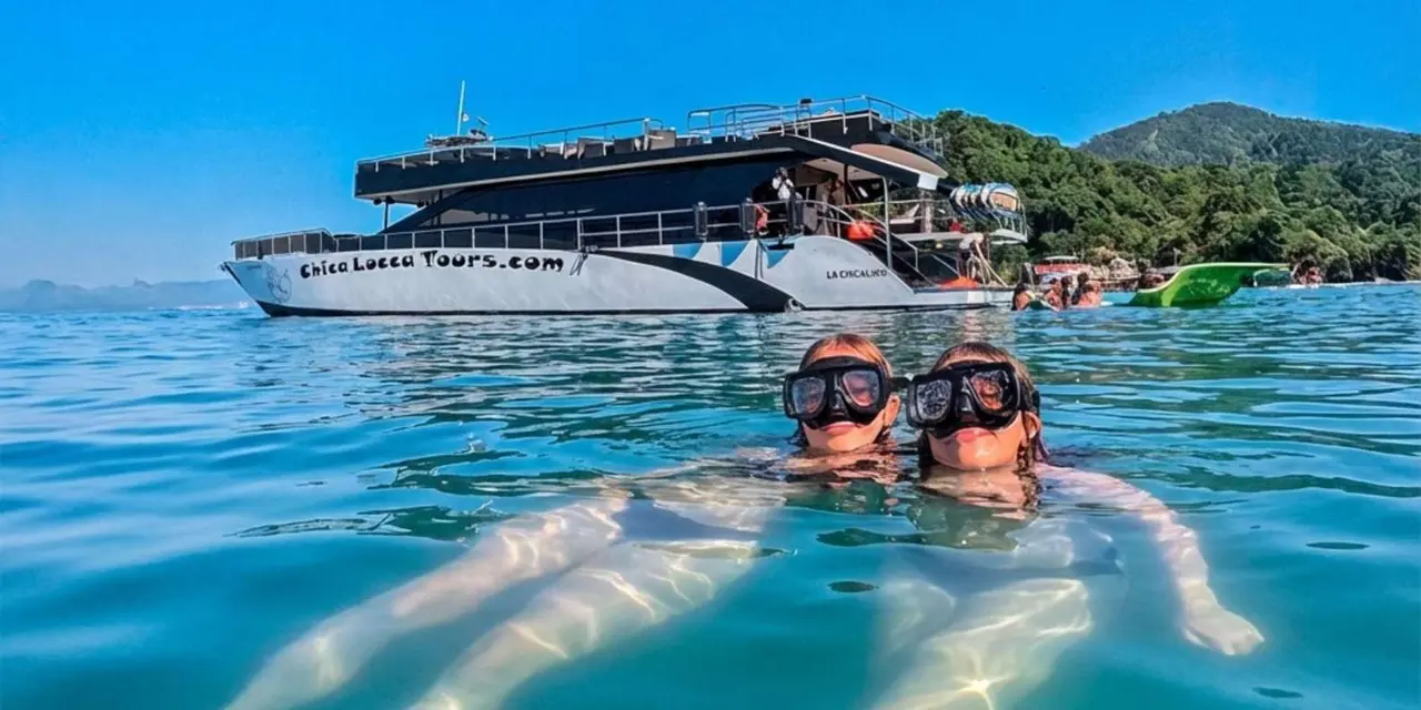 Two snorkelers in masks floating in clear water in front of a large white catamaran with a sun deck