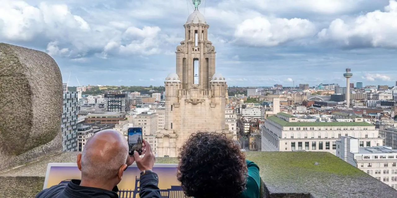 Two visitors photographing the domed tower of the Royal Liver Building and the city skyline from a stone rooftop viewing platform