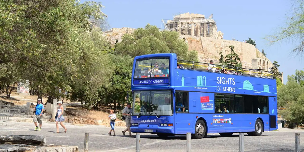 A blue open-top Sights of Athens double-decker bus, the Parthenon on the Acropolis above the trees behind it