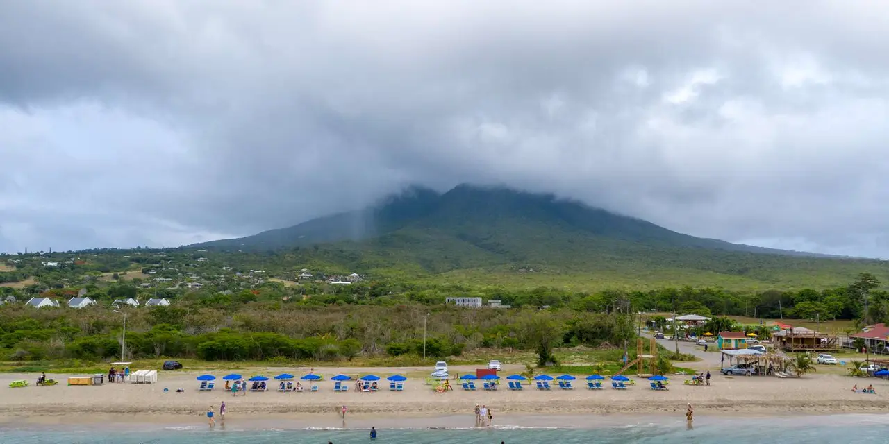 Rows of blue beach umbrellas on a sandy Nevis beach under a cloud-capped Nevis Peak