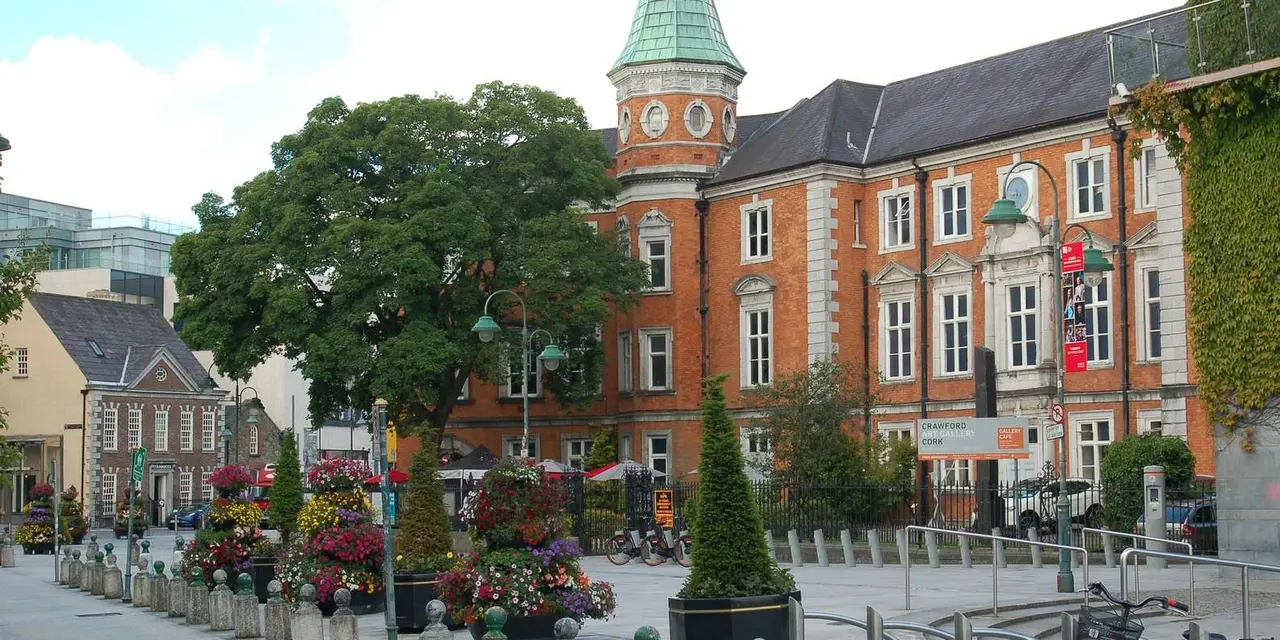 A red-brick building with a green copper-roofed clock tower above a flower-lined street in Cork