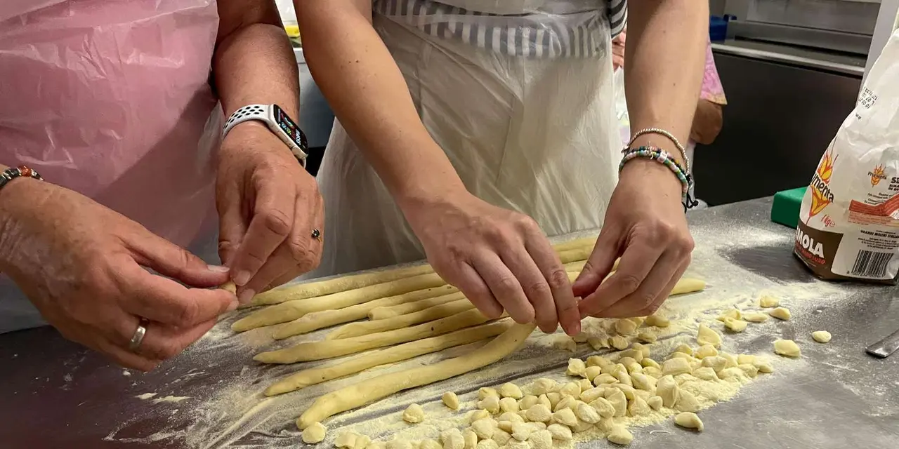 Two pairs of hands rolling and cutting fresh pasta dough on a floured steel counter