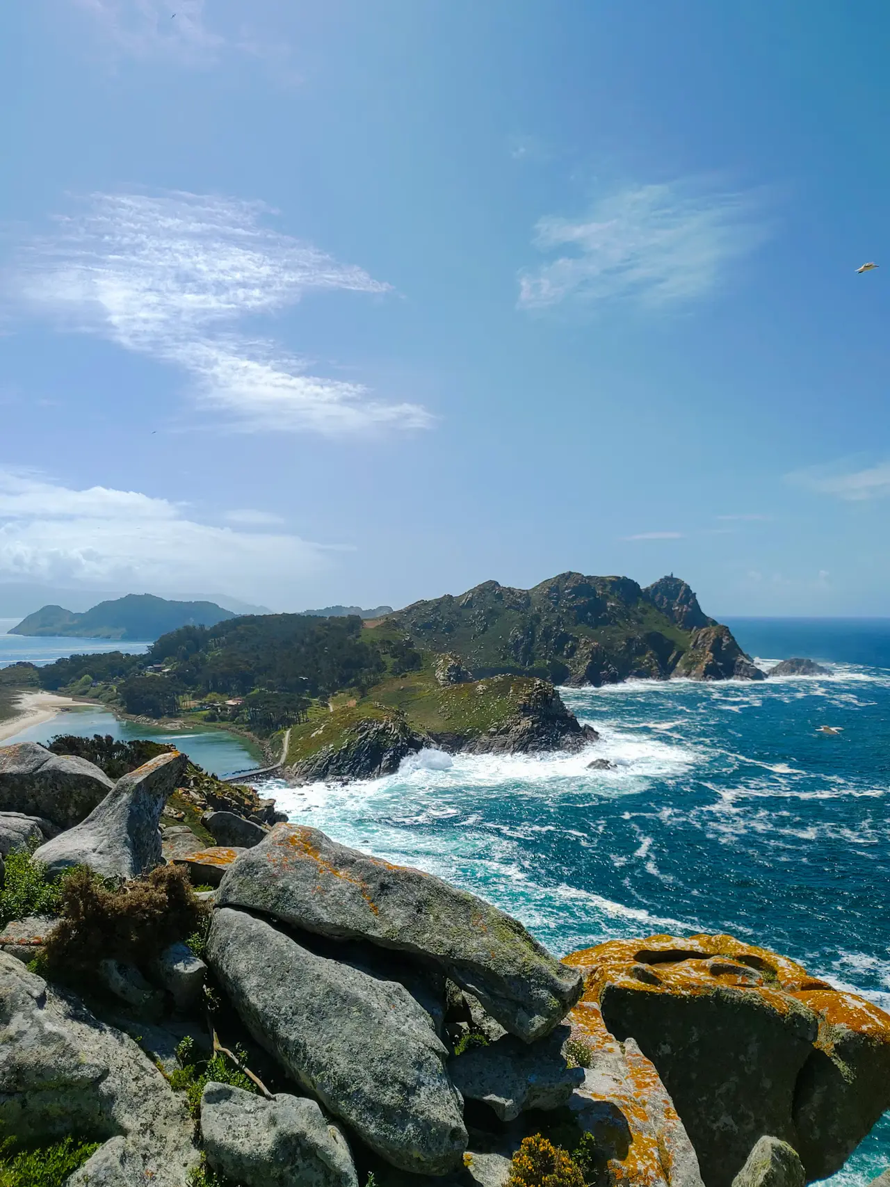 The Cíes Islands, at the mouth of the Ría de Vigo, seen from a rocky summit on Monteagudo: green, craggy headlands, white surf on a deep-blue Atlantic and a pale sand beach on the calm side, under a wide blue sky