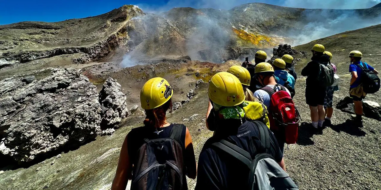 Trekkers in yellow helmets and backpacks walking beside a steaming volcanic crater