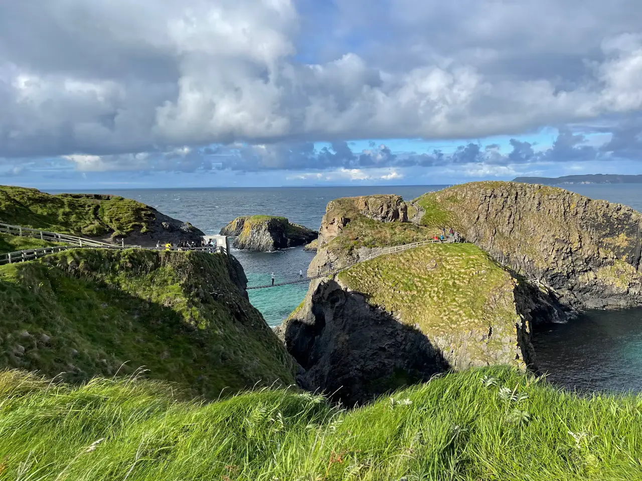 A rope bridge stretching between two green sea stacks above turquoise water, with grey clouds and open sea behind