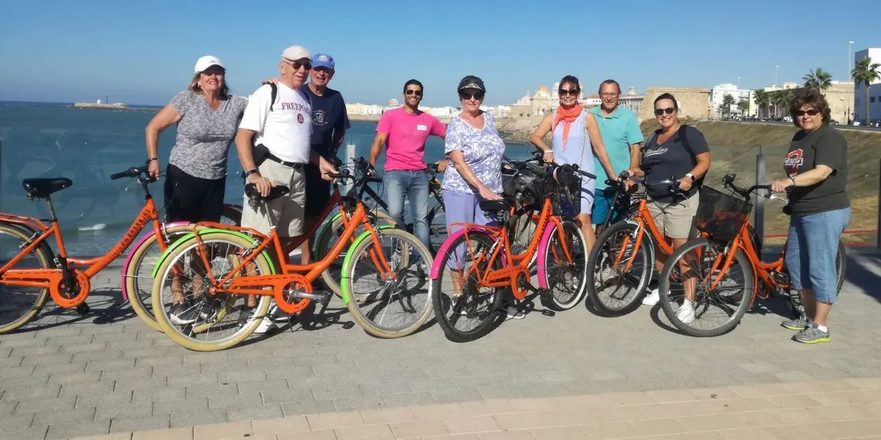 A group of eight people posing with orange rental bikes on a waterfront promenade with the sea behind them