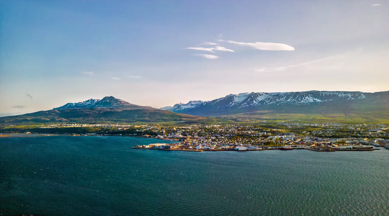Akureyri seen across Eyjafjörður at golden hour, with the town along the waterfront and snow-streaked mountains behind under a pale blue sky