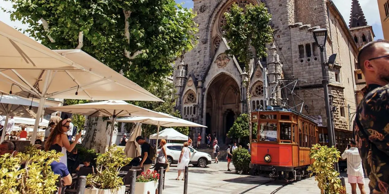 A red vintage tram on a street beside a stone church in Sóller with café umbrellas