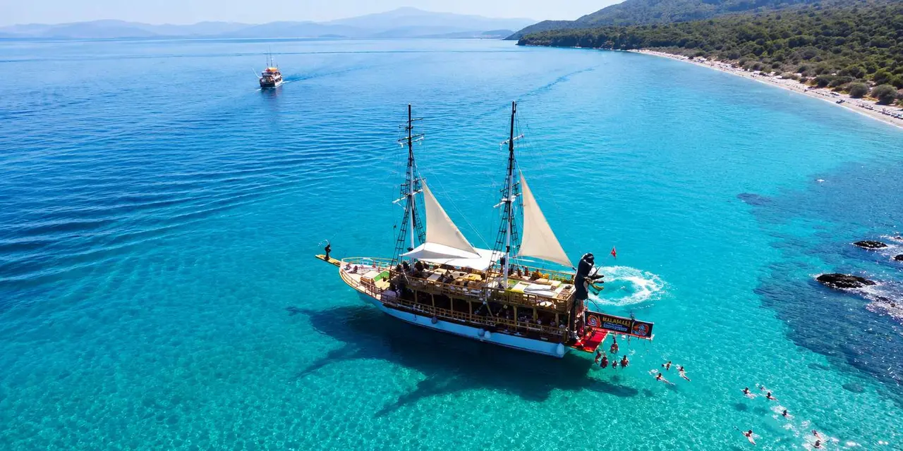 A two-masted wooden tour boat on turquoise water with swimmers around it and a forested headland behind