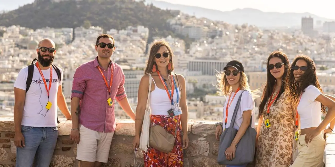 A group of smiling visitors wearing lanyards on a terrace, the white buildings of Athens spreading over the hills behind