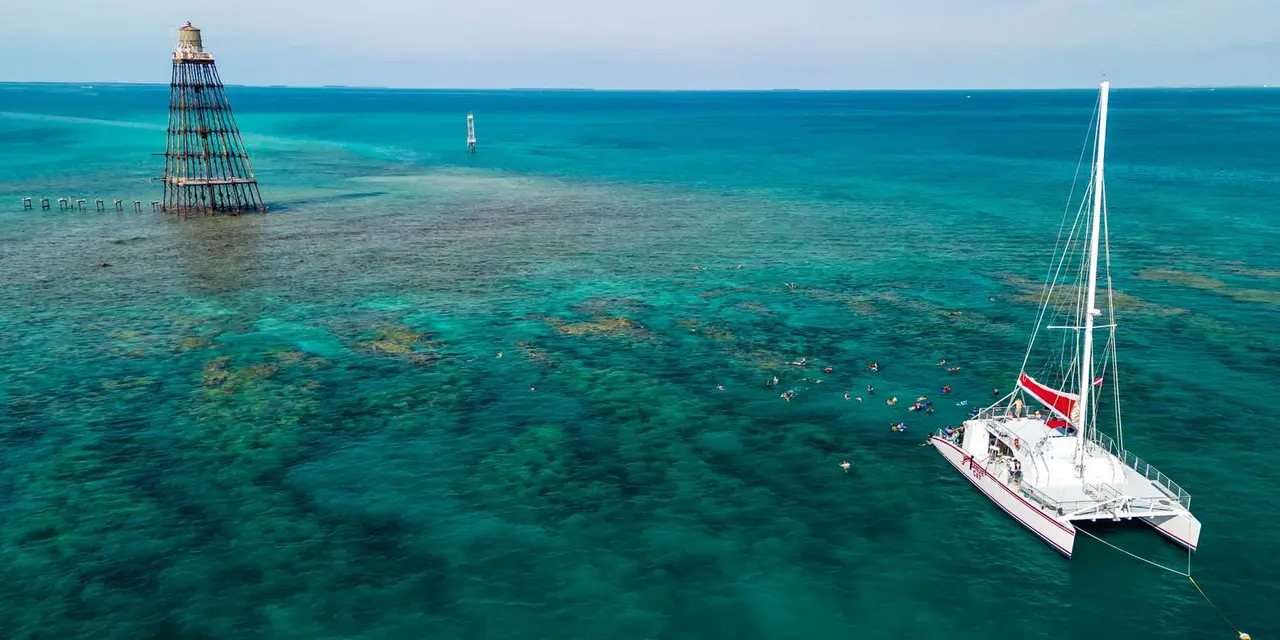 An aerial view of a white catamaran moored beside a rusting offshore lighthouse tower, with snorkelers spread across the turquoise reef