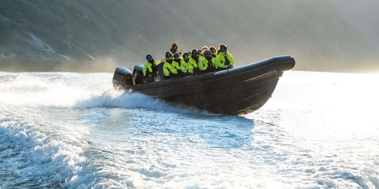 A dark inflatable boat full of passengers in yellow suits speeding across a sunlit fjord