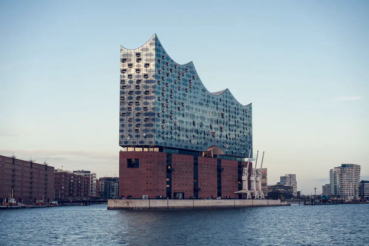 The glass-topped Elbphilharmonie concert hall on its red-brick base, seen across the Elbe at Hamburg's HafenCity under a pale blue evening sky
