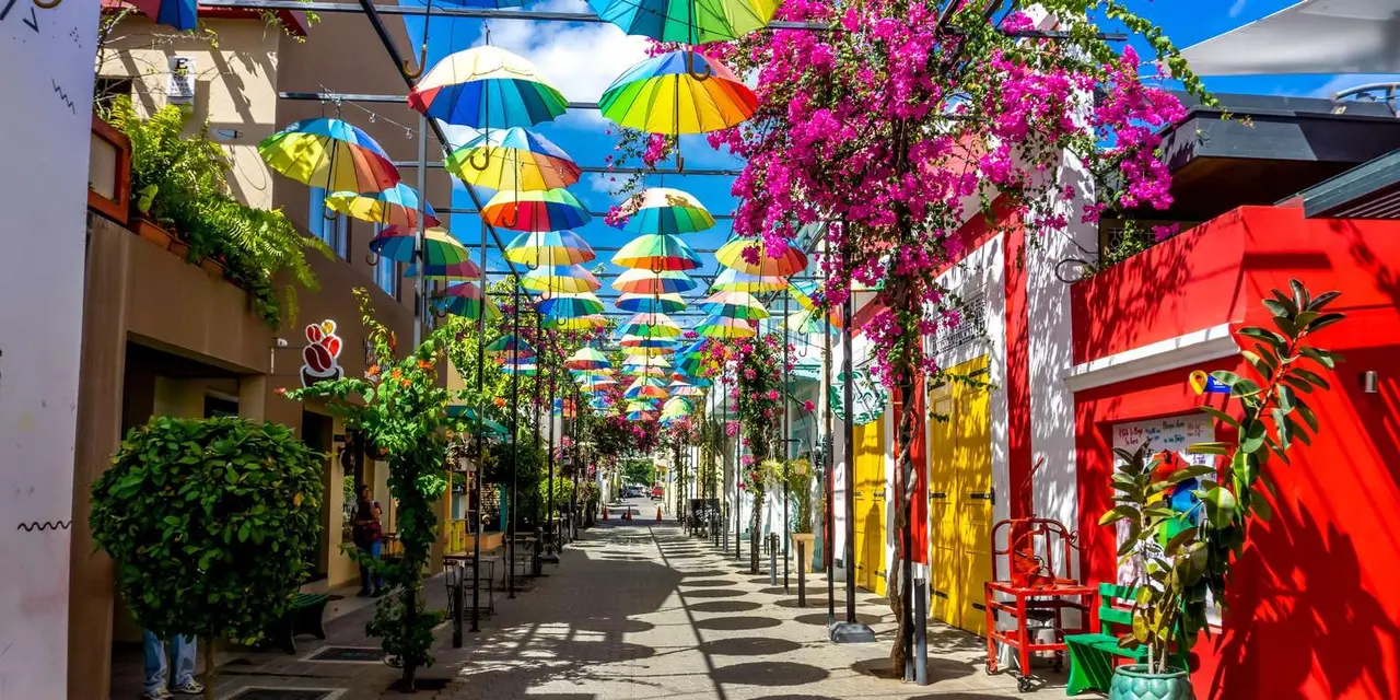 Rainbow umbrellas hanging over a cobbled street lined with bougainvillea and brightly painted shopfronts