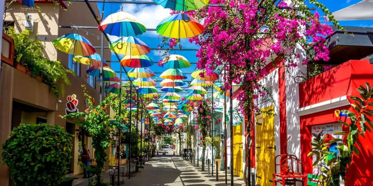 Rainbow umbrellas hanging over a cobbled street lined with bougainvillea and brightly painted shopfronts