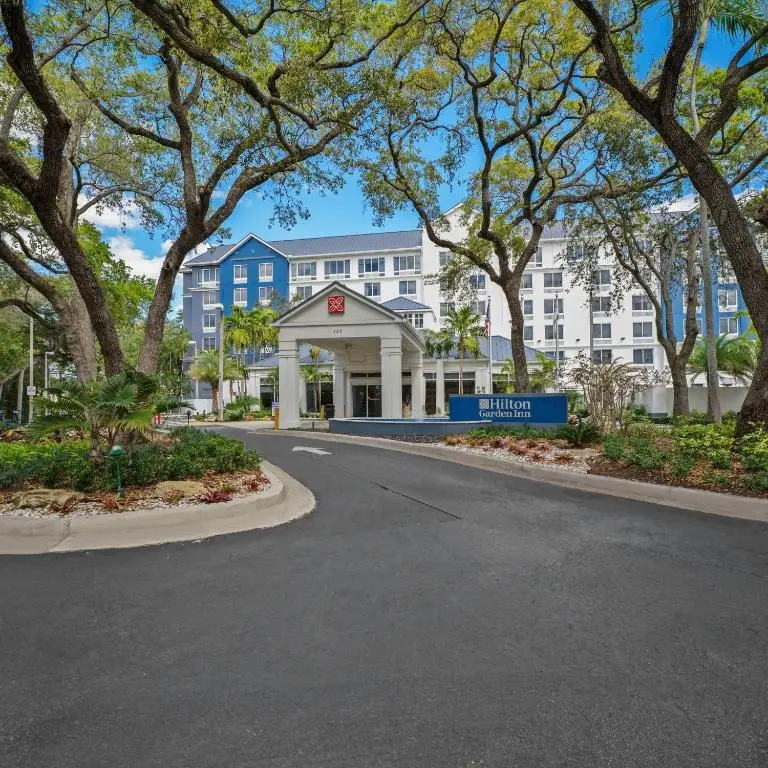 The blue-and-white Hilton Garden Inn Ft. Lauderdale Airport-Cruise Port behind a covered entrance and oak trees