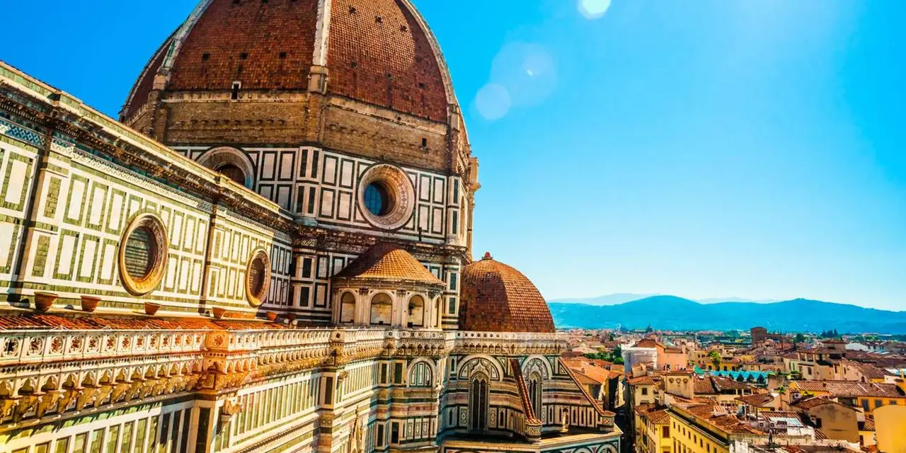 A close view of Florence cathedral's red dome and its pink, green and white marble walls against a bright blue sky
