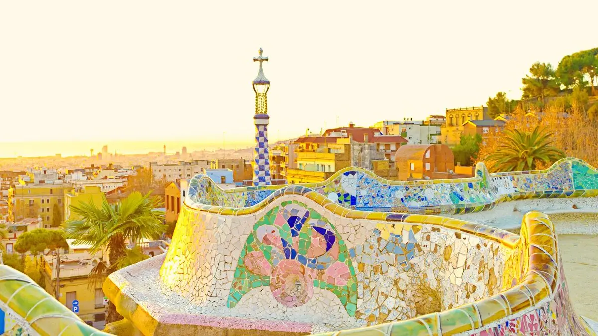 Park Güell's curving mosaic bench in warm light, with a tiled spire and the rooftops of Barcelona fading toward the sea
