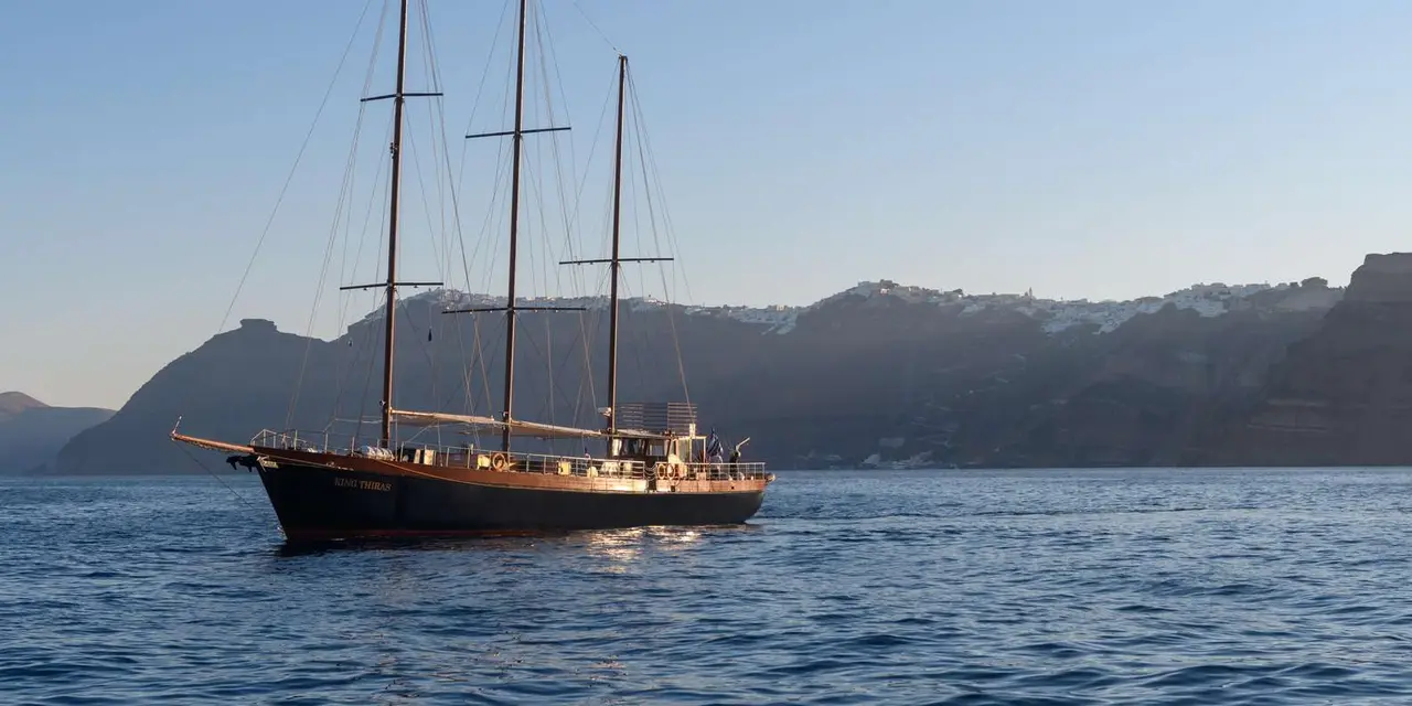 The three-masted wooden sailing ship King Thiras on calm water with white villages on the caldera rim behind it